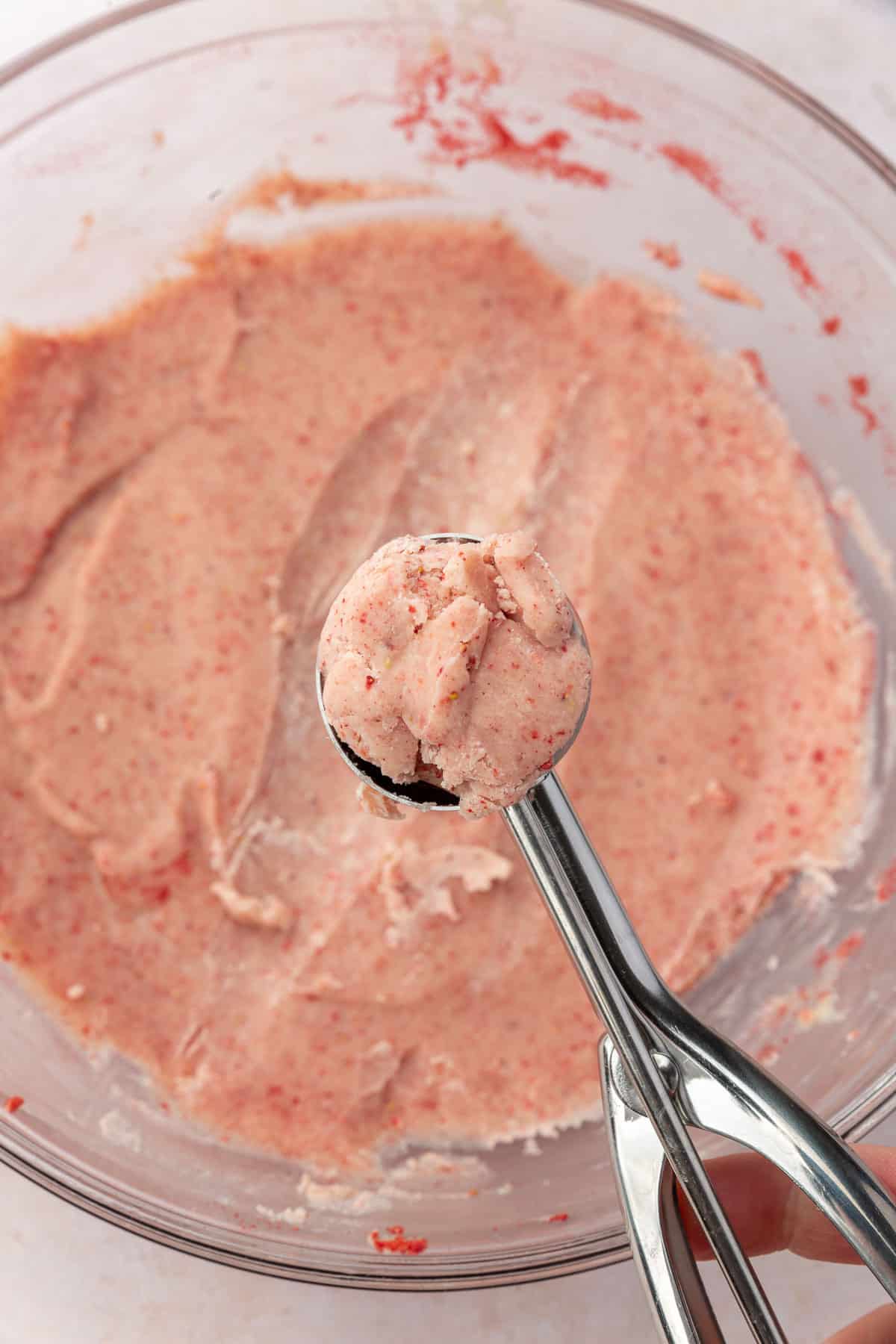 A stainless steel scooper filled with strawberry truffle over a glass bowl of more strawberry white chocolate truffle dough.