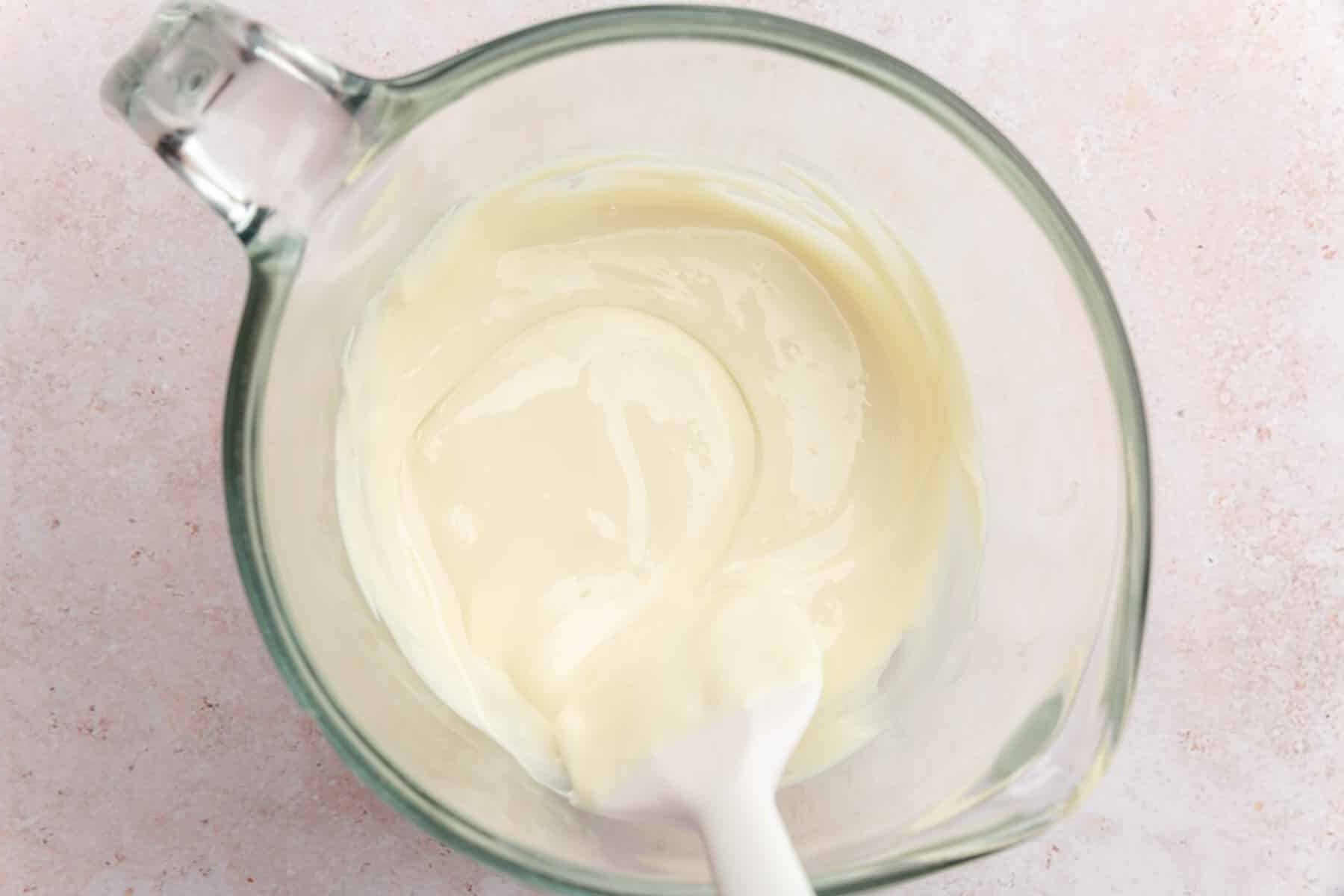 A measuring cup with melted white chocolate and a light pink spatula.