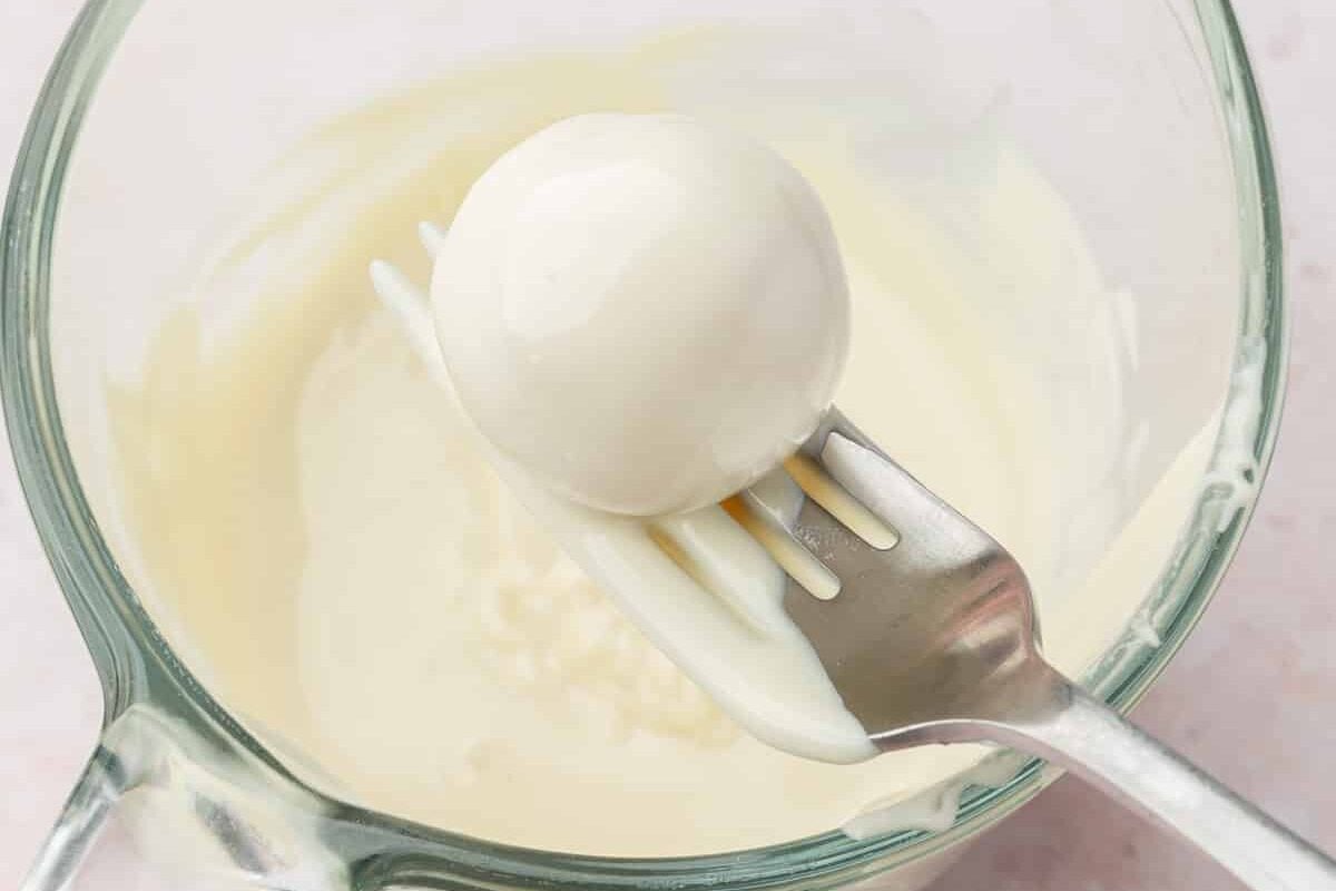 A fork dipping a strawberry truffle into a measuring cup of melted white chocolate.