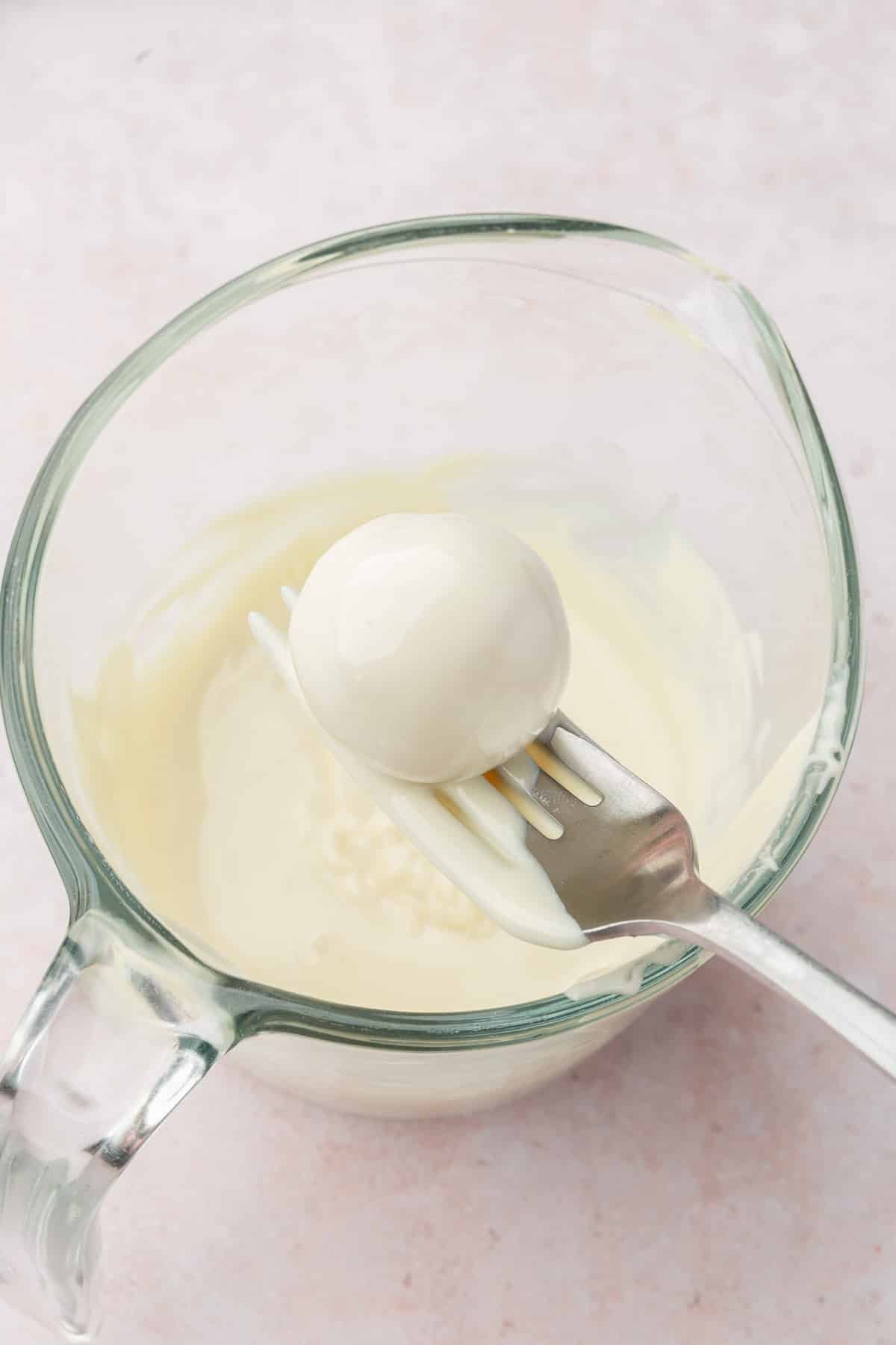 A fork dipping a strawberry truffle into a measuring cup of melted white chocolate.