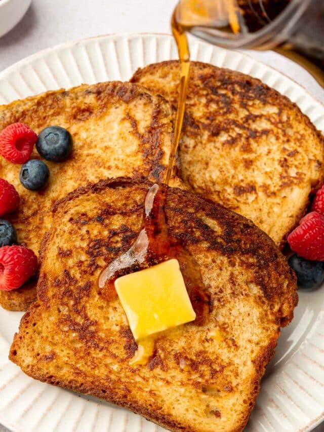 A small pitcher of maple syrup being poured over three slices of gluten-free french toast topped with melty butter and fresh berries.