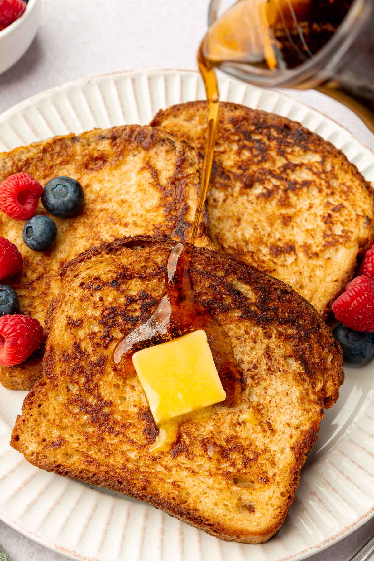 A small pitcher of maple syrup being poured over three slices of gluten-free french toast topped with melty butter and fresh berries.