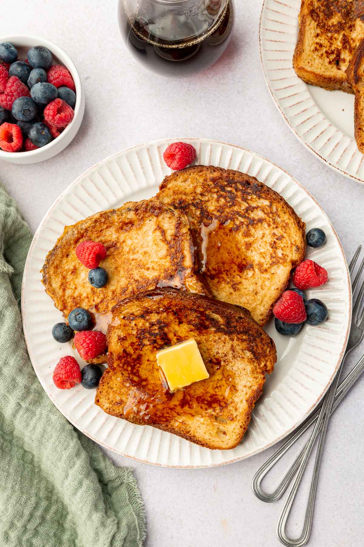 A plate with three slices of gluten-free french toast topped with butter, maple syrup, blueberries and raspberries with additional french toast and berries to the side.