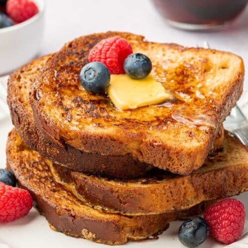 A stack of four slices of gluten-free french toast topped with fresh blueberries, raspberries, butter and maple syrup.