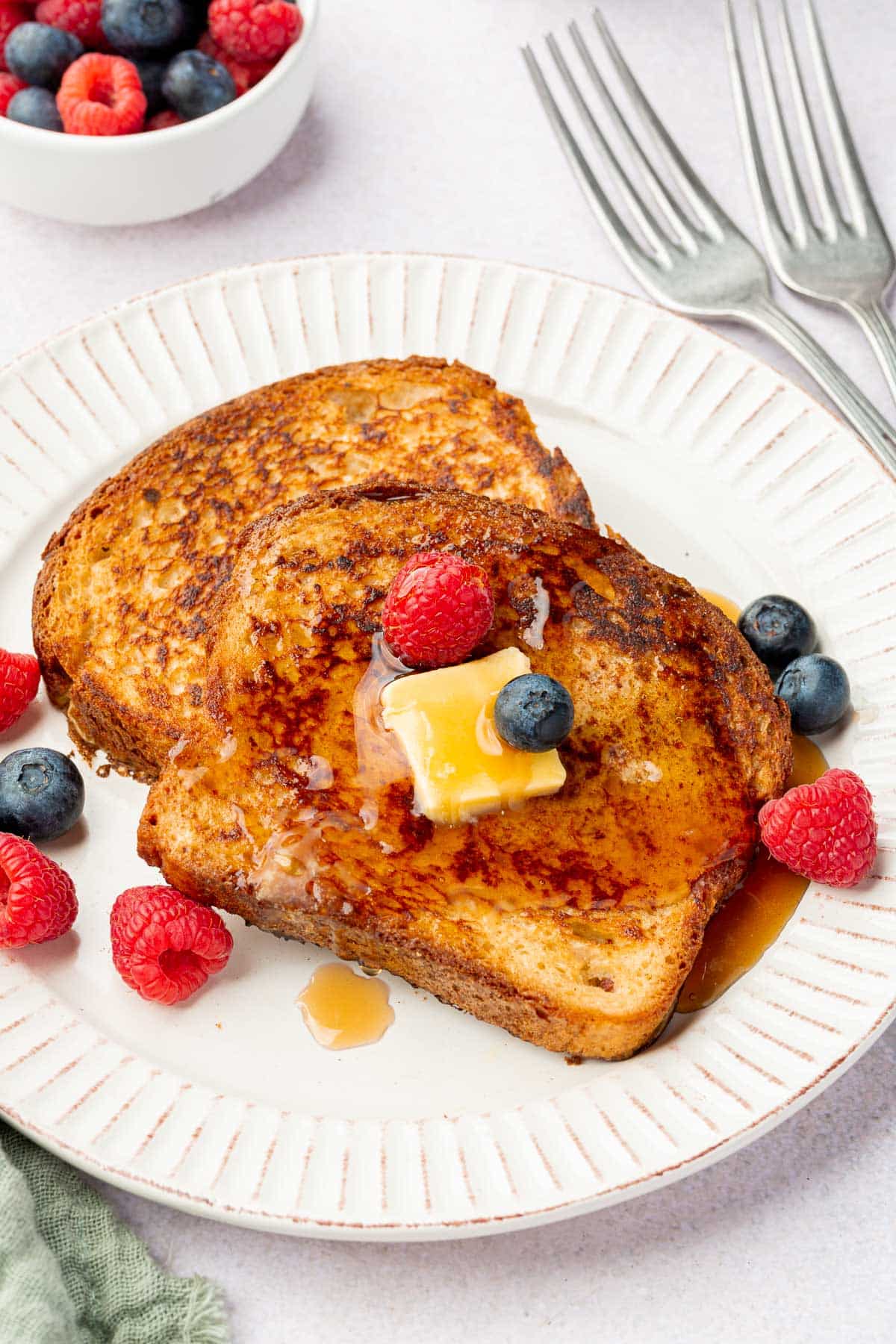 A white plate with two pieces of gluten-free french toast topped with a pat of butter, maple syrup and fresh raspberries and blueberries.