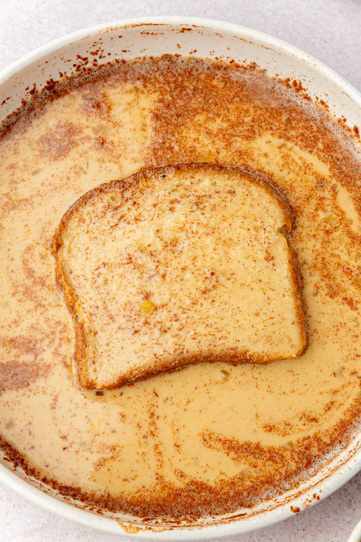 A shallow bowl with a cinnamon and egg mixture that has a piece of gluten-free bread soaking in it.