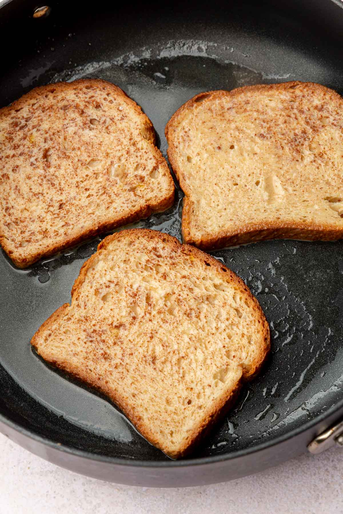 A non-stick skillet with three slices of gluten-free french toast that are cooking in melted butter.