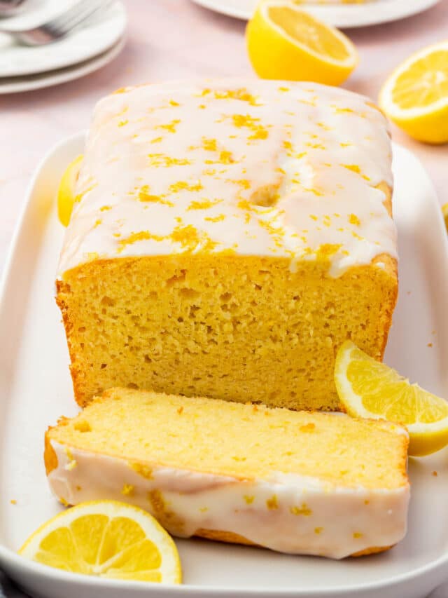 A gluten free lemon drizzle loaf cake that has the front piece sliced off on a platter with fresh lemons on the surface.