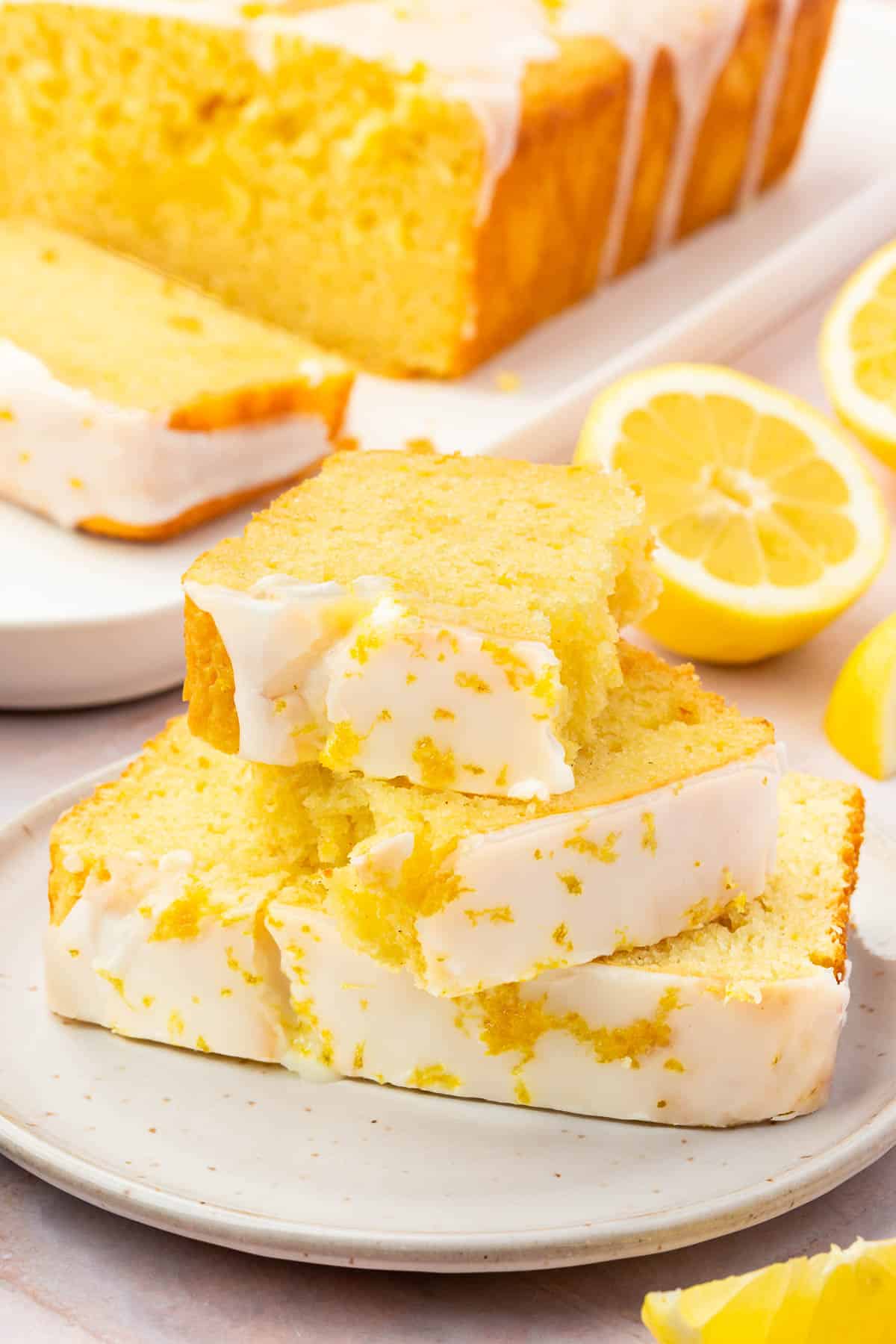 A stack of lemon drizzle cake slices on a dessert plate with the rest of the lemon cake on a platter in the background.