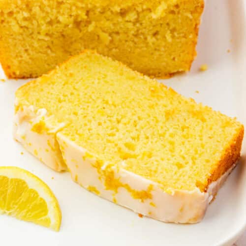 A slice of a gluten free lemon loaf with lemon glaze on a white platter with additional cake behind it and some fresh lemon slices.