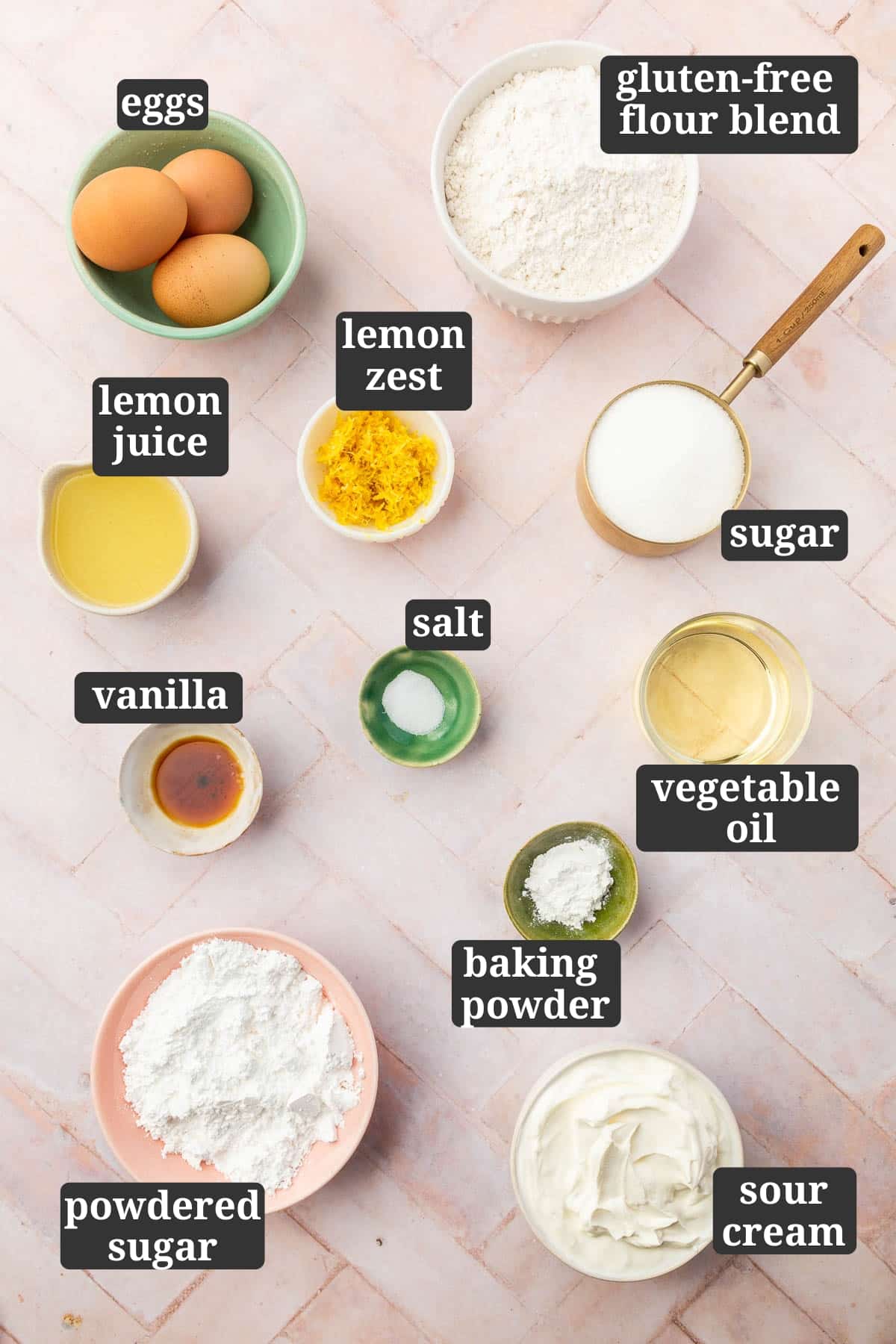 An overhead view of ingredients in small bowls to make gluten-free lemon drizzle cake, including, eggs, gluten-free flour, lemon juice, lemon zest, sugar, vanilla, salt, vegetable oil, baking powder, powdered sugar and sour cream with text overlays over each ingredient.
