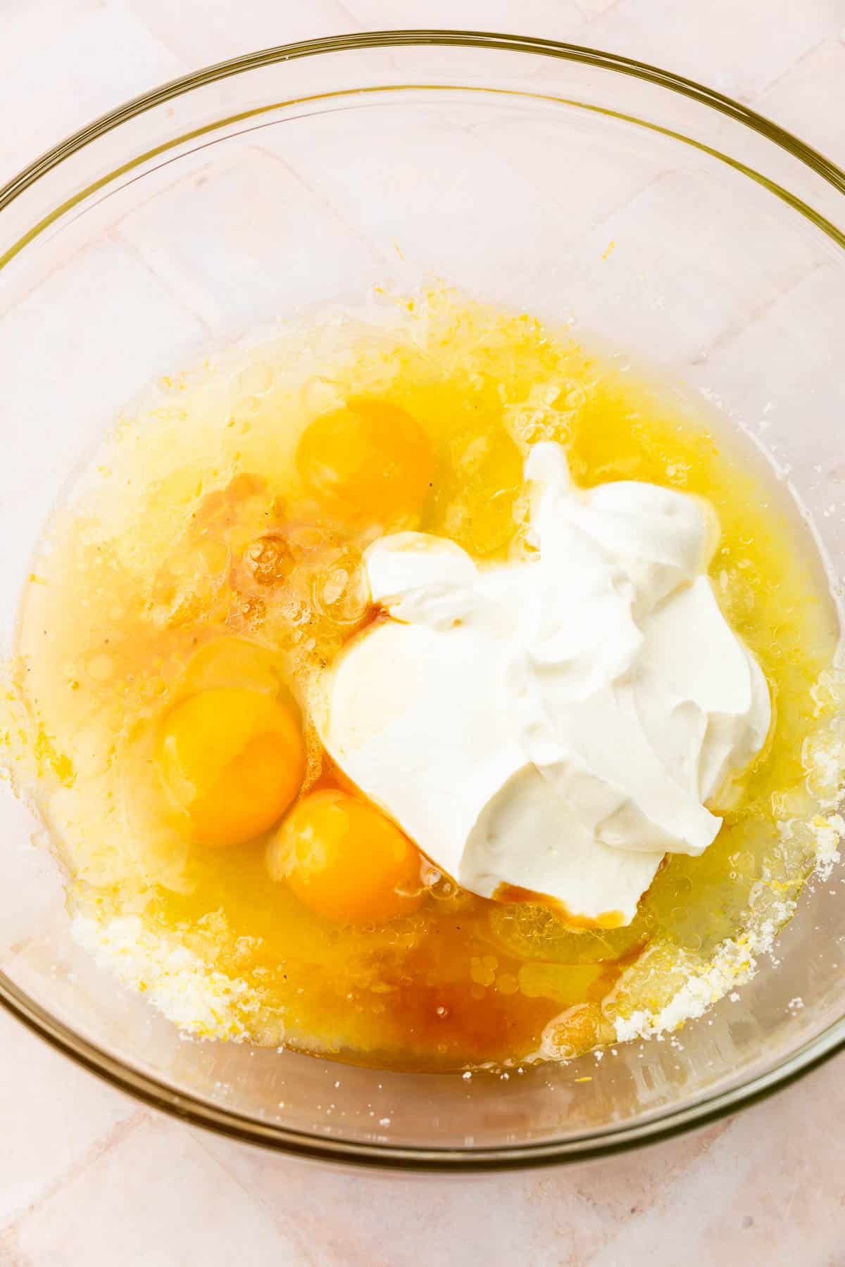 A glass mixing bowl with lemon zest, granulated sugar, lemon juice, 3 eggs, sour cream, and vanilla in it before mixing together.