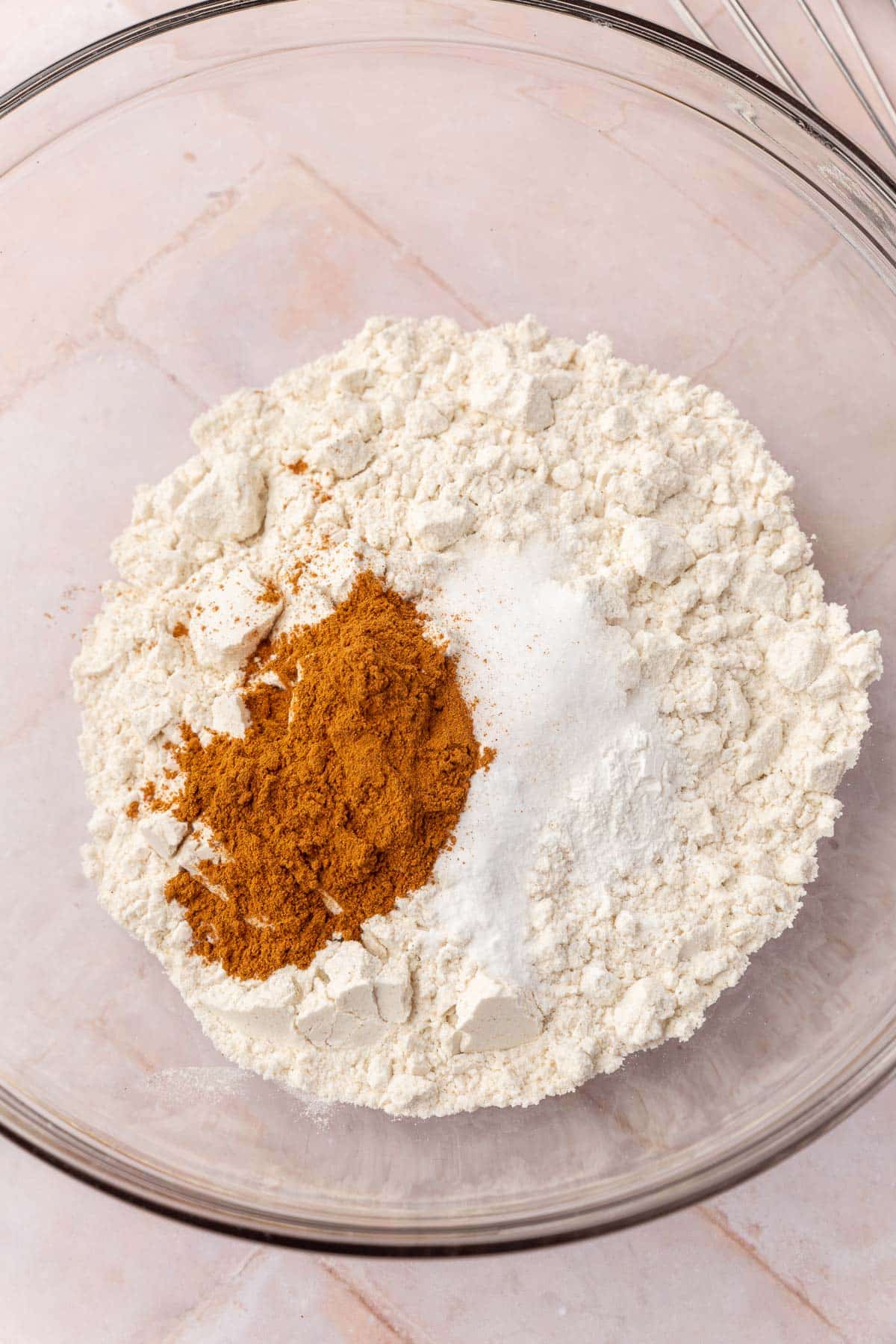 A glass mixing bowl with gluten-free flour, ground cinnamon, salt, baking soda and baking powder before whisking together.