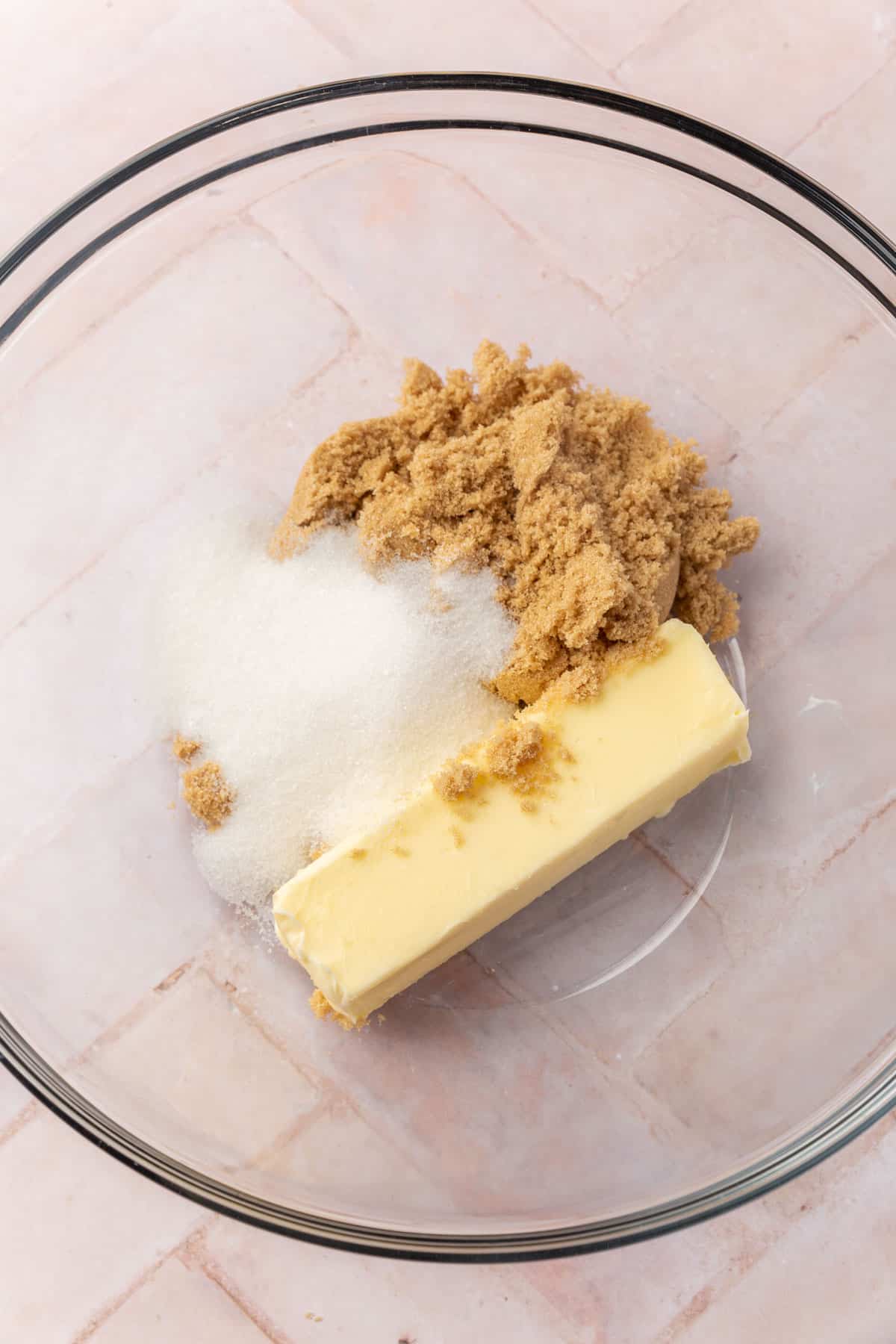 A glass mixing bowl with a stick of butter, brown sugar, and granulated sugar.