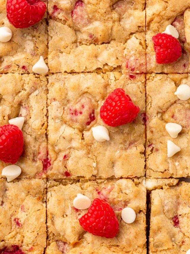 A close up of a slab of gf white chocolate raspberry blondies cut into 9 equal squares with fresh raspberries and white chocolate chips decorating the top.