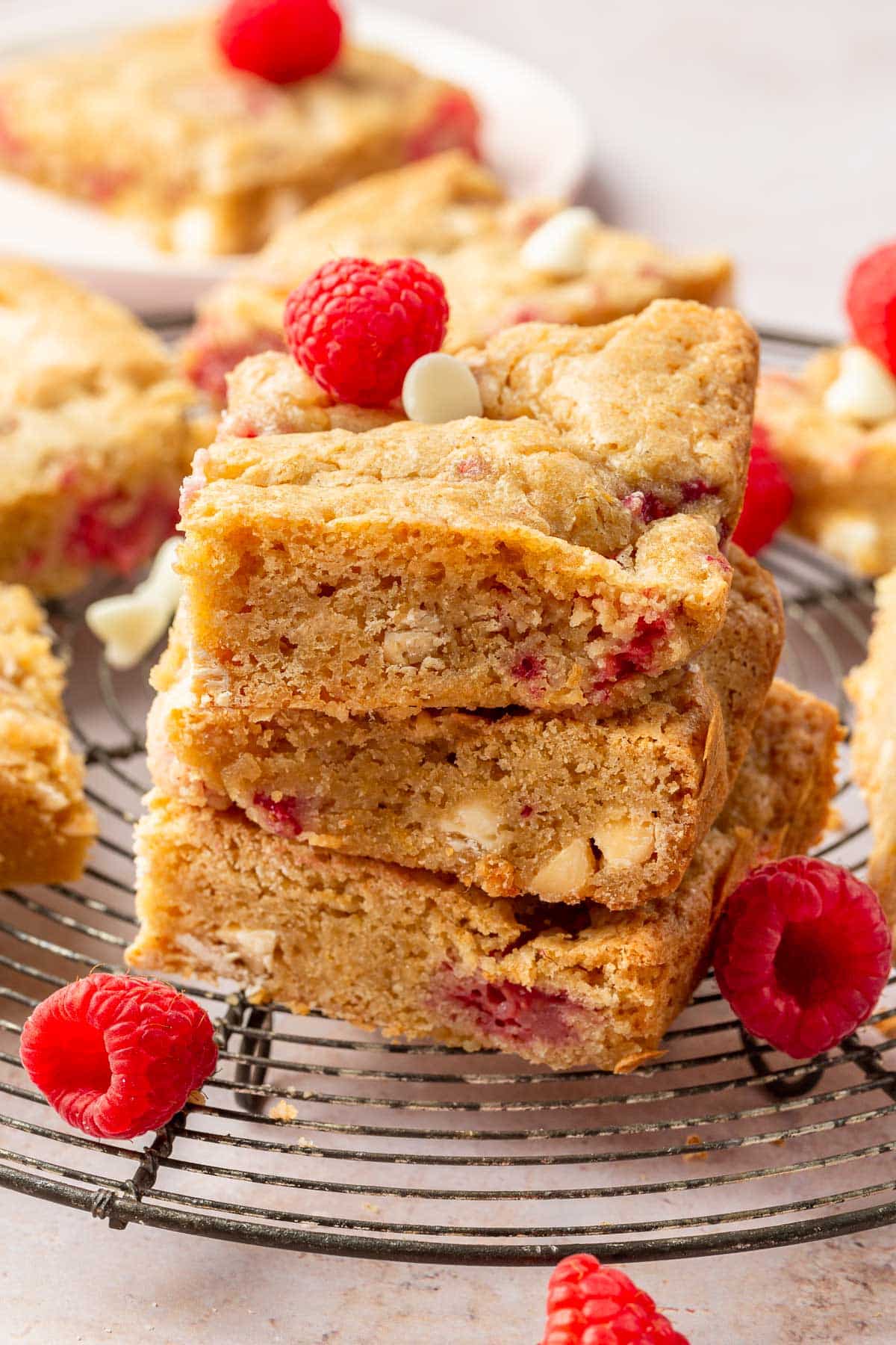 A stack of three gluten-free raspberry white chocolate blondies on a cooling rack surrounded by fresh raspberries.