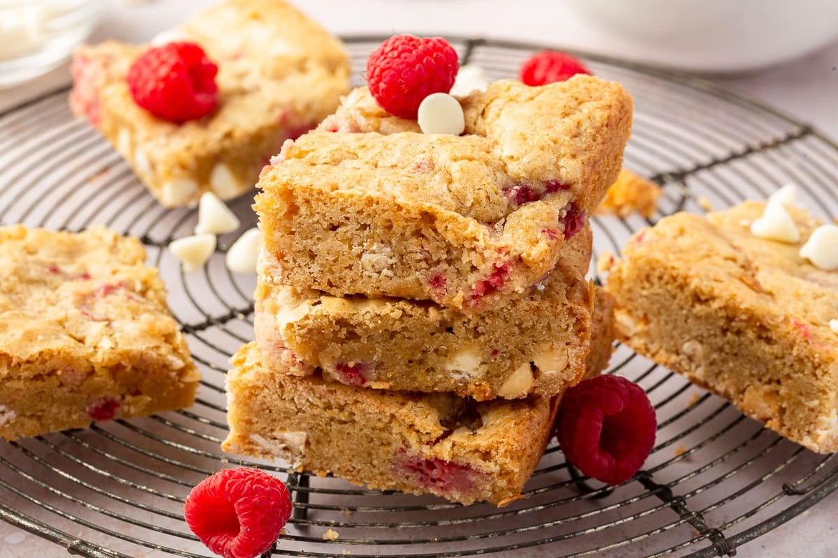 A stack of three gf raspberry white chocolate blondies on a wire cooling rack with additional blondies topped with raspberries and white chocolate chips.