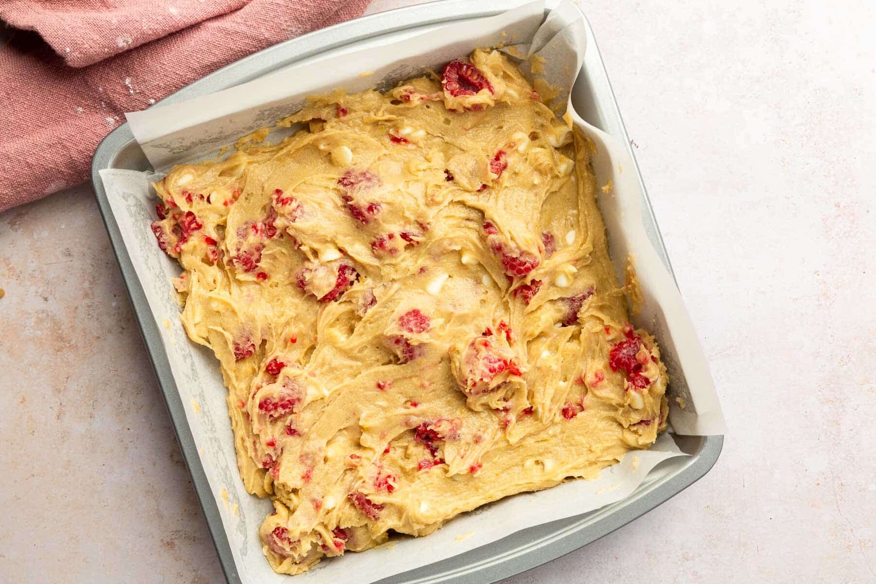 A square metal baking dish lined with parchment paper that has gluten free raspberry white chocolate blondie batter in it before baking in the oven.