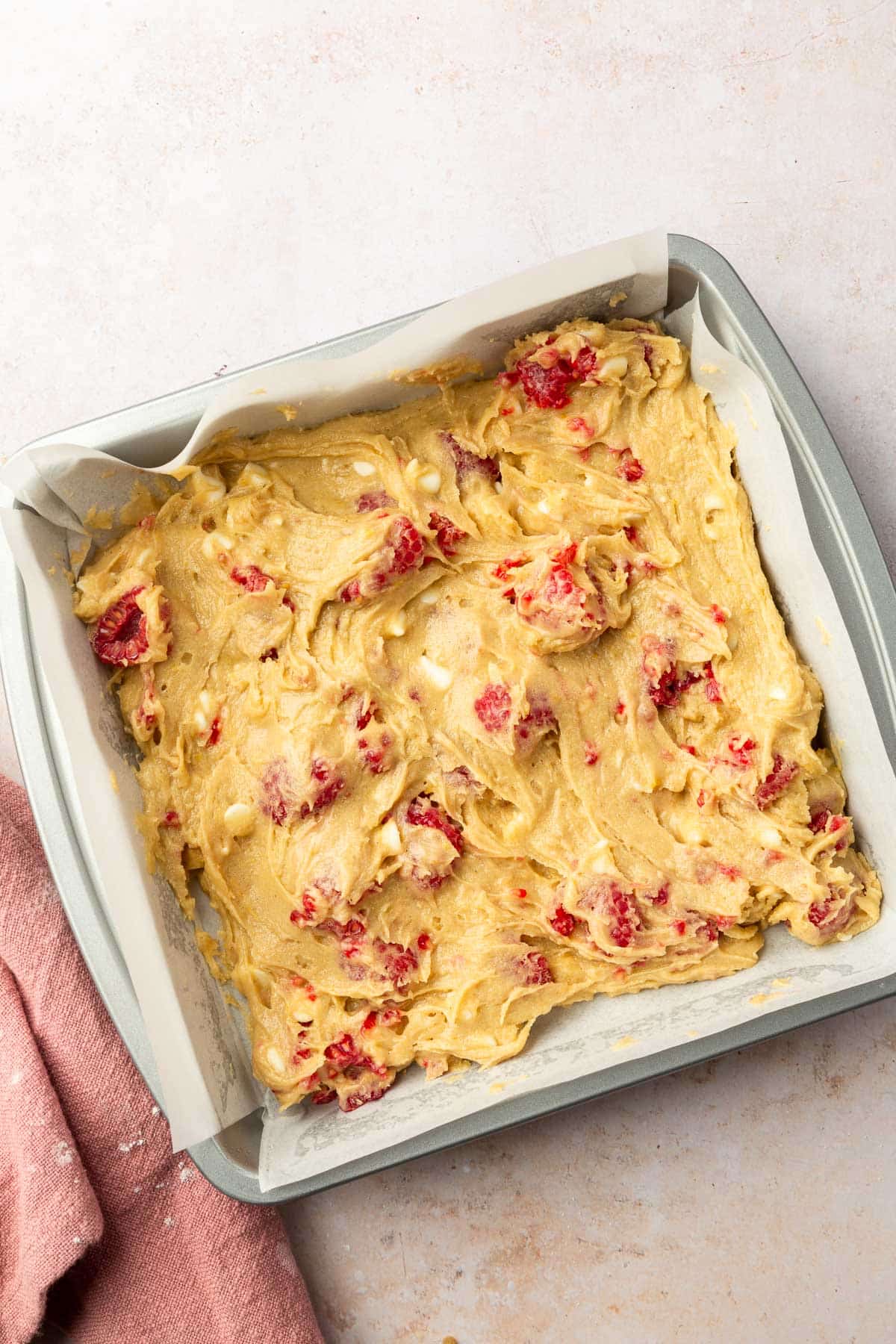 A square metal baking dish lined with parchment paper that has gluten free raspberry white chocolate blondie batter in it before baking in the oven.