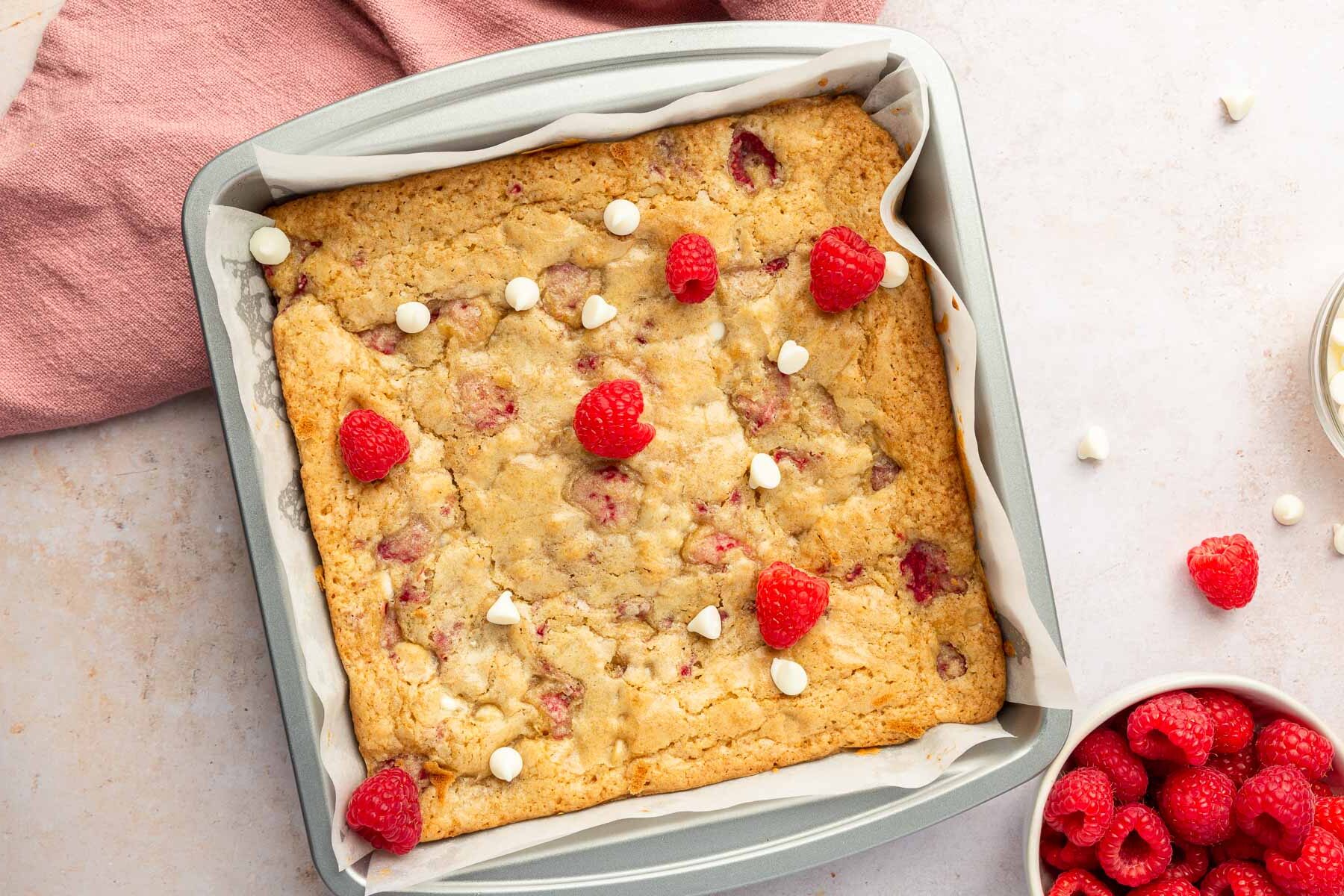 A square metal baking pan filled with gluten free raspberry white chocolate blondies that have been baked in the oven with a bowl of raspberries to the side.
