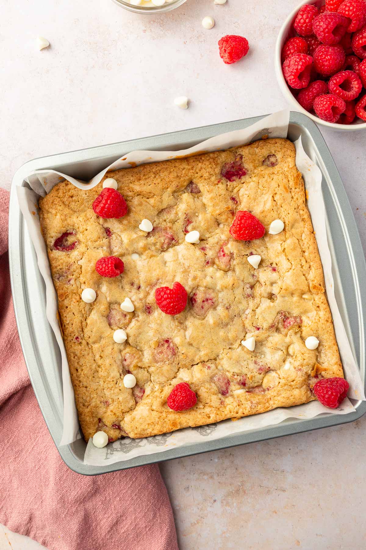 A square metal baking pan filled with gluten free raspberry white chocolate blondies that have been baked in the oven with a bowl of raspberries to the side.