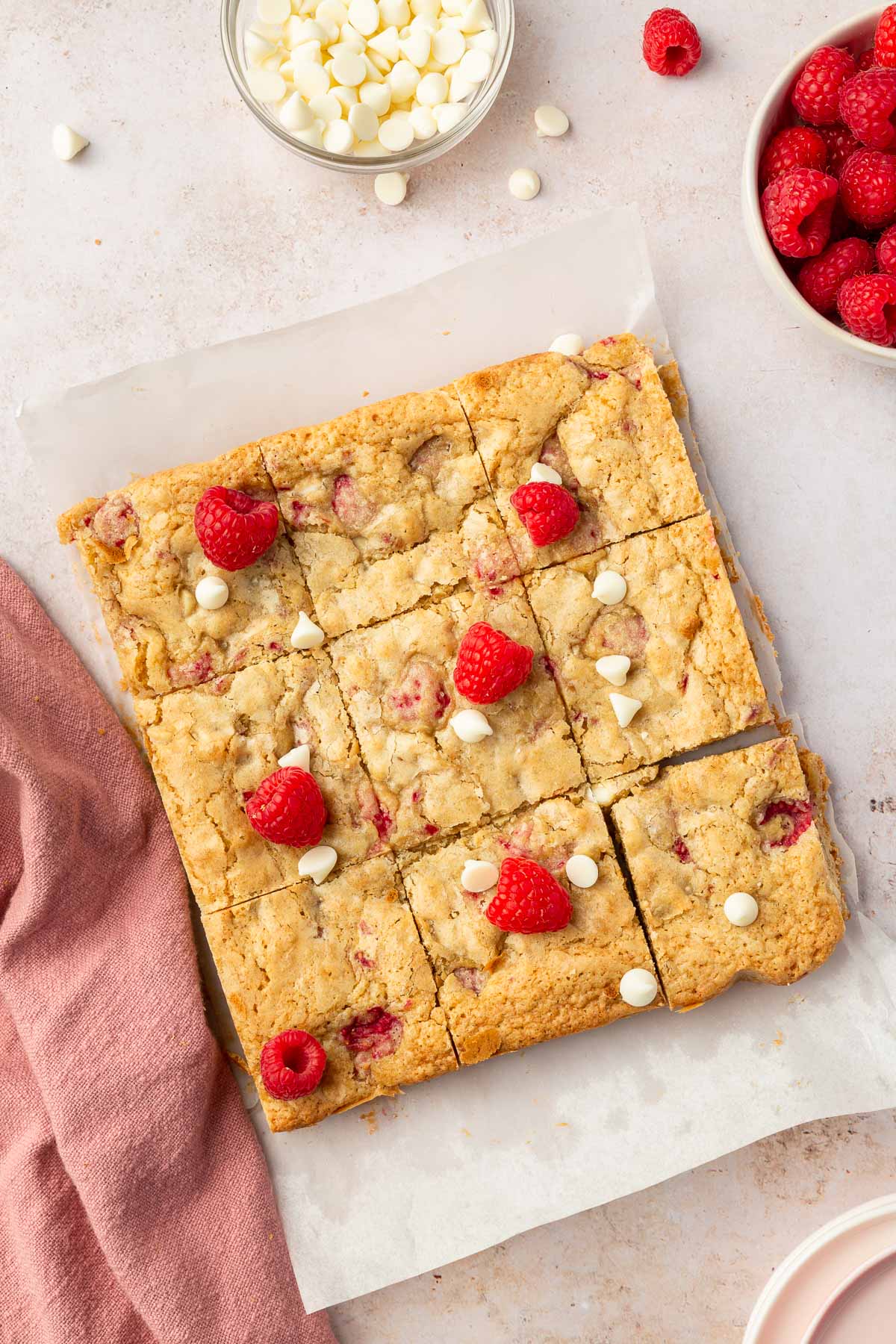 An overhead view of a slab of white chocolate raspberry blondies cut into 9 equal sections on a piece of parchment paper and decorated with fresh raspberries.