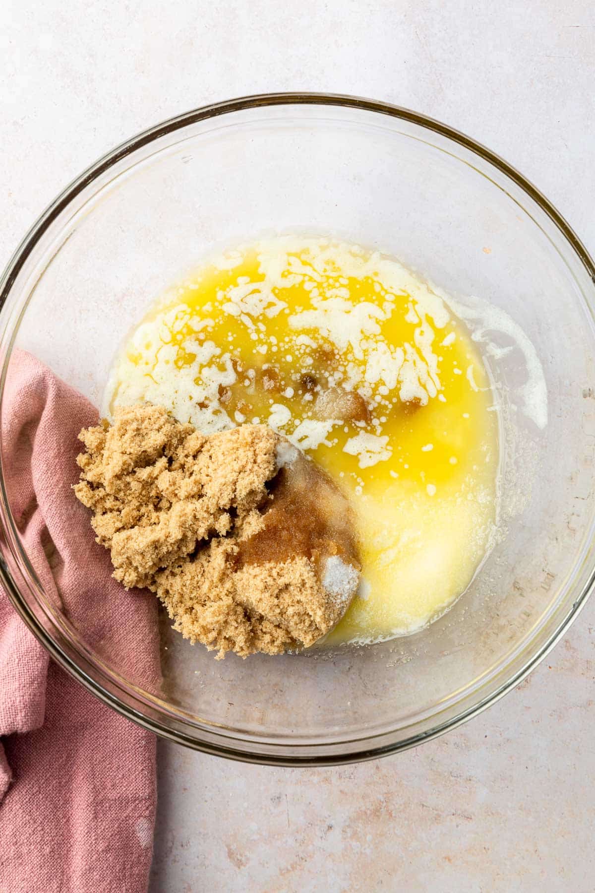 A glass mixing bowl with brown sugar, granulated sugar and melted butter in it before mixing together.