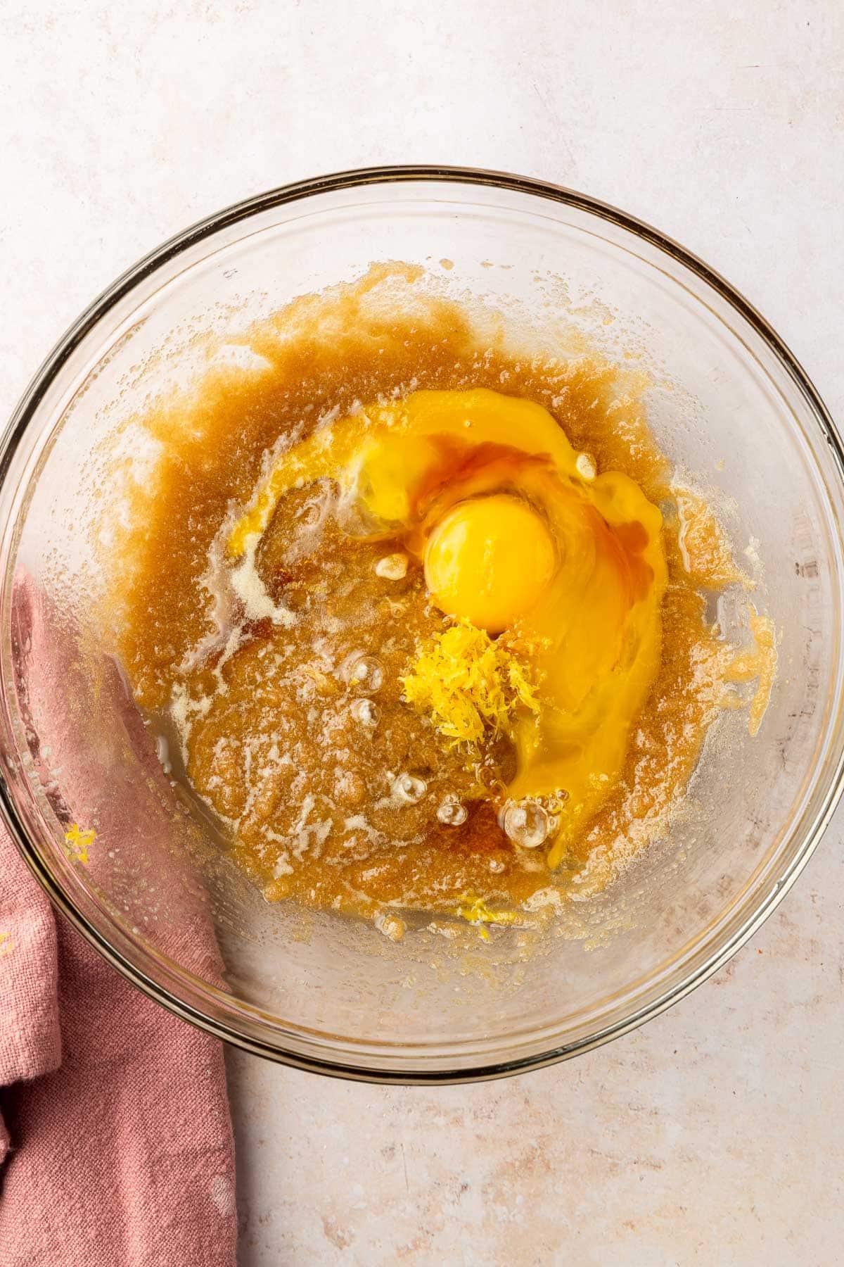 A glass mixing bowl with brown sugar and melted butter mixture topped with an egg, egg yolk, lemon zest, and vanilla extract.