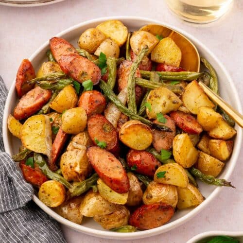 A bowl of roasted potatoes, sausage, green beans and onions with a bowl of fresh parsley and a glass of wine to the side.