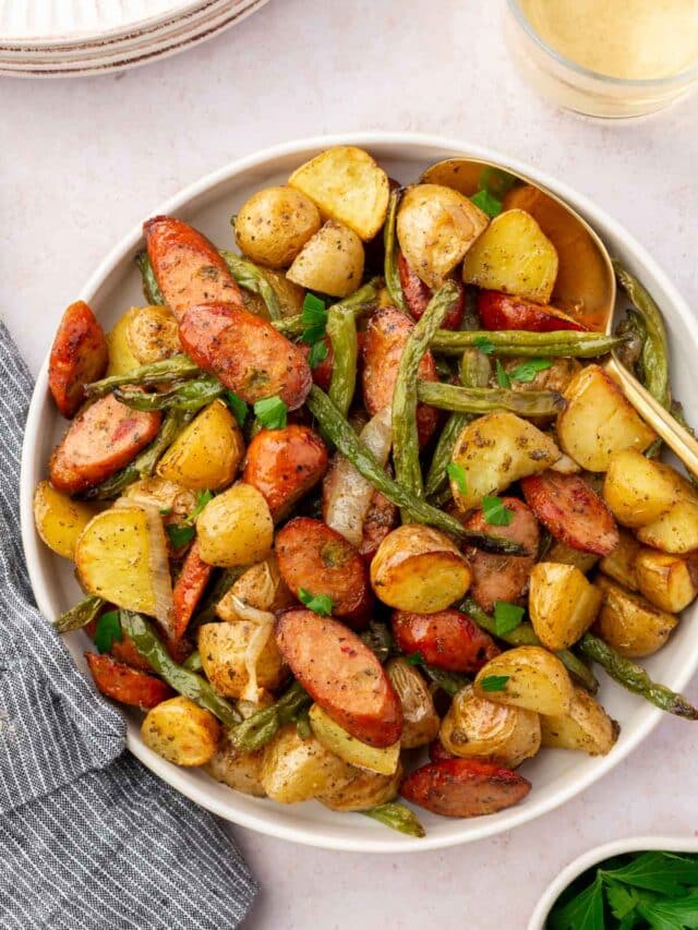 An overhead view of roasted green beans, potatoes, sausage and onions in a bowl with a gold serving spoon.