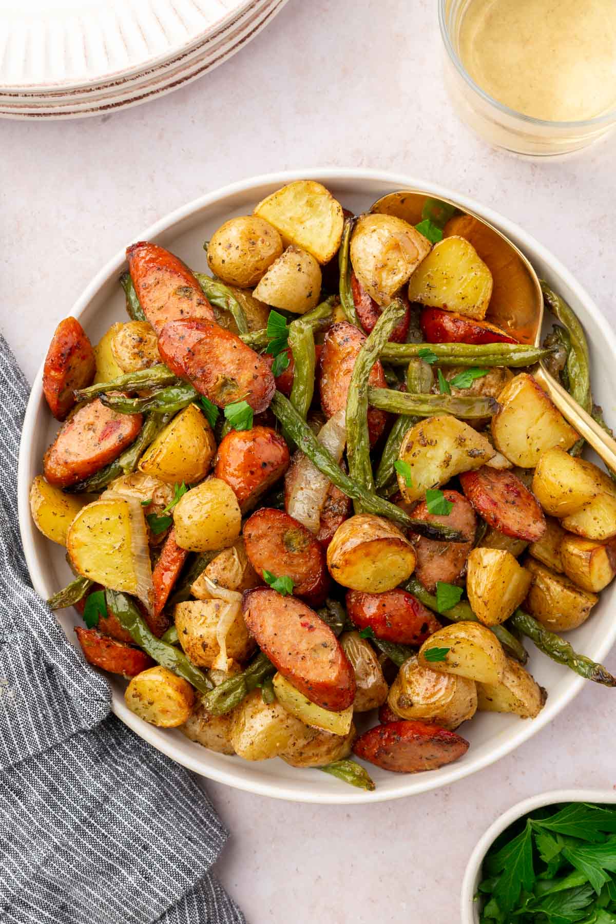 An overhead view of roasted green beans, potatoes, sausage and onions in a bowl with a gold serving spoon.