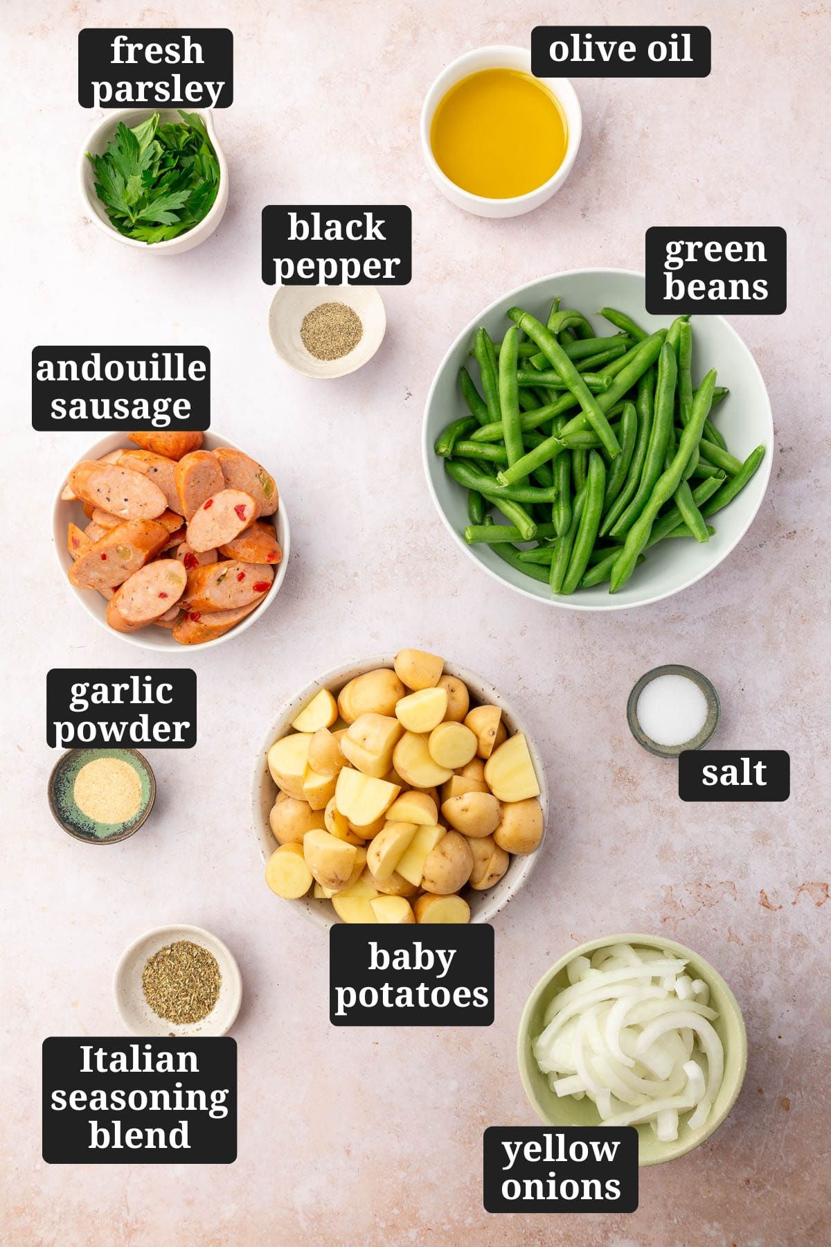 An overhead view of ingredients in bowls to make sheet pan sausage, green beans, and potatoes, including fresh parsley, black pepper, olive oil, green beans, andouille sausage, garlic powder, baby potatoes, salt, Italian seasoning blend, and yellow onions with text overlays over each ingredient.