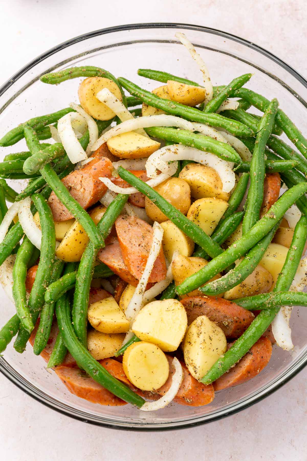 a glass bowl filled with green beans, sausage, onions and potatoes tossed in oil and spices.