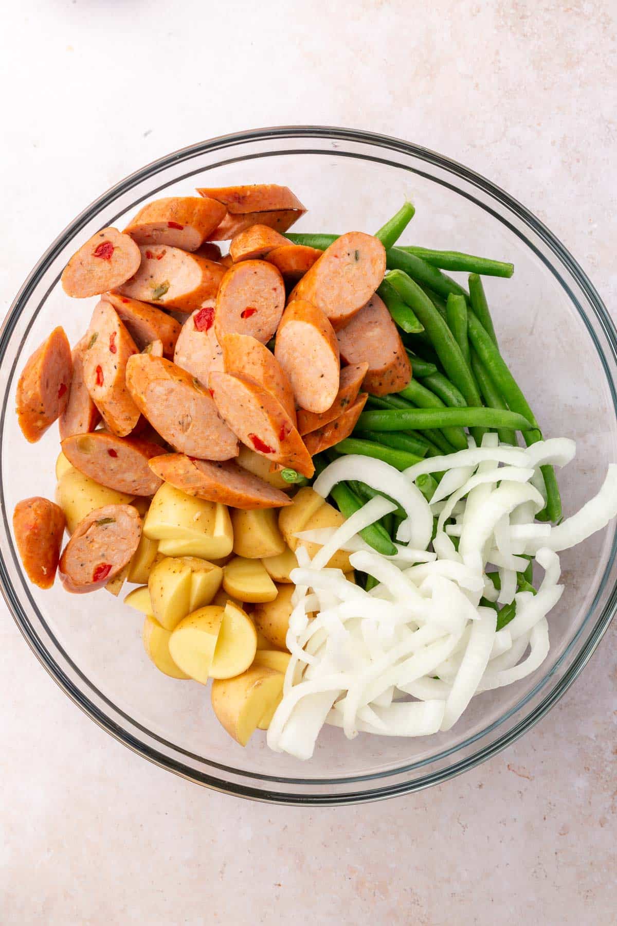 A glass mixing bowl of andouille sausage, green beans, yellow onions, and baby potatoes before mixing together.