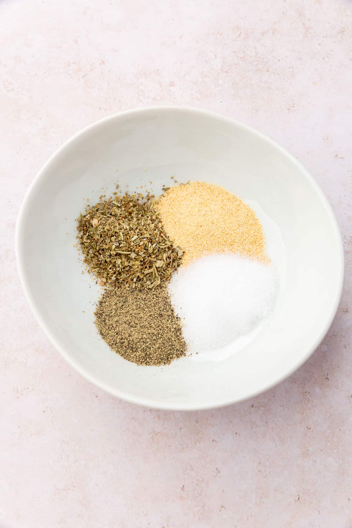 A small white bowl of Italian seasoning blend, garlic powder, salt, and black pepper that have not been mixed together.