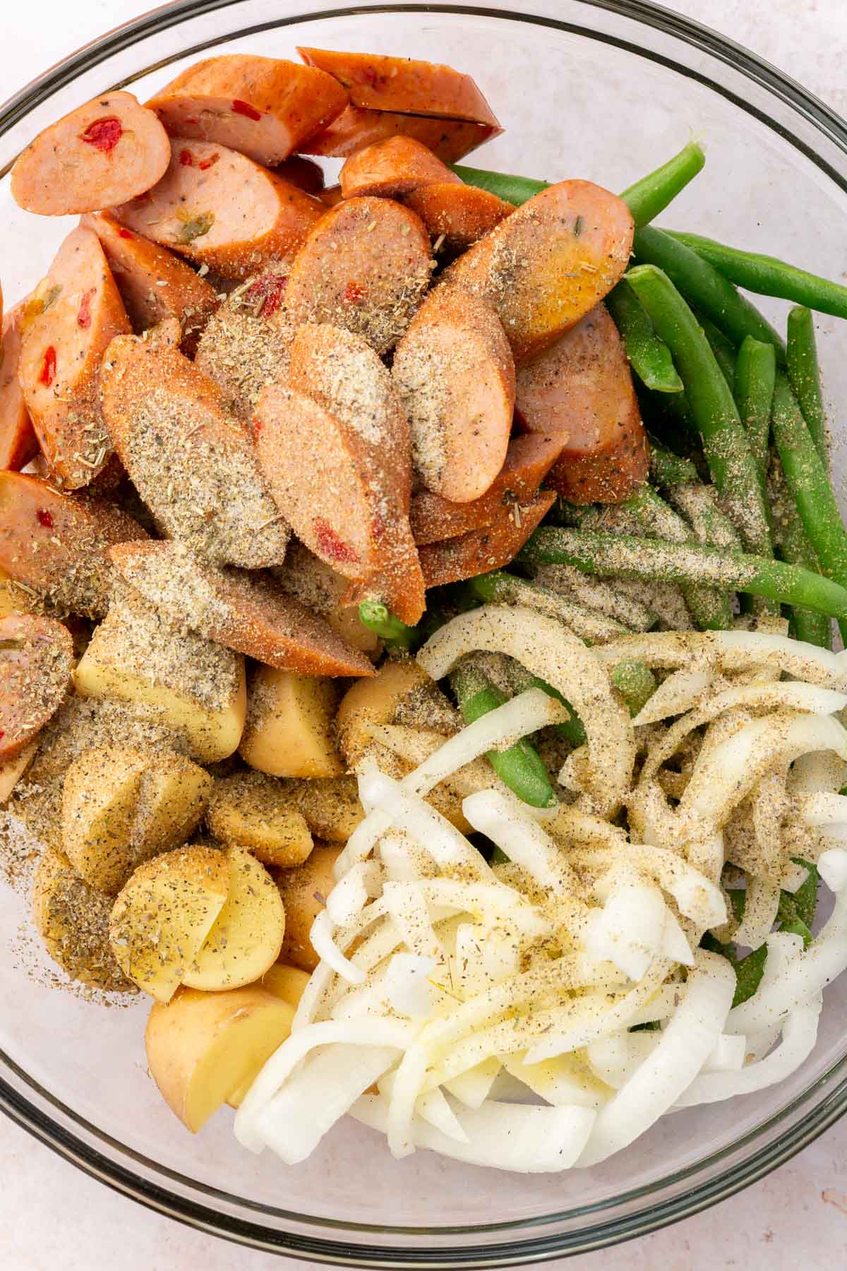 A glass bowl filled with sliced sausage, onions, green beans, potatoes topped with dried spices.