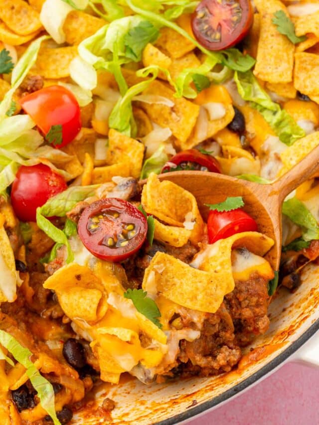 A skillet filled with Frito taco casserole with ground beef, tomatoes and lettuce and wood spoon scooping a portion out of the skillet.