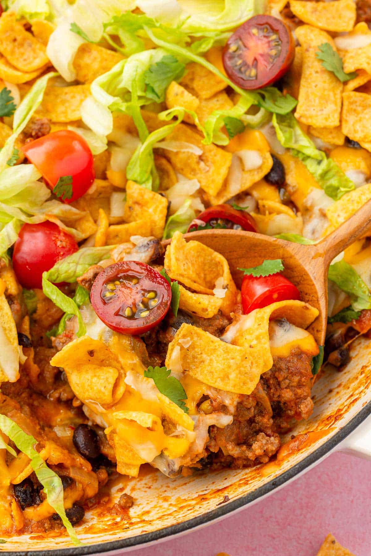 A skillet filled with Frito taco casserole with ground beef, tomatoes and lettuce and wood spoon scooping a portion out of the skillet.