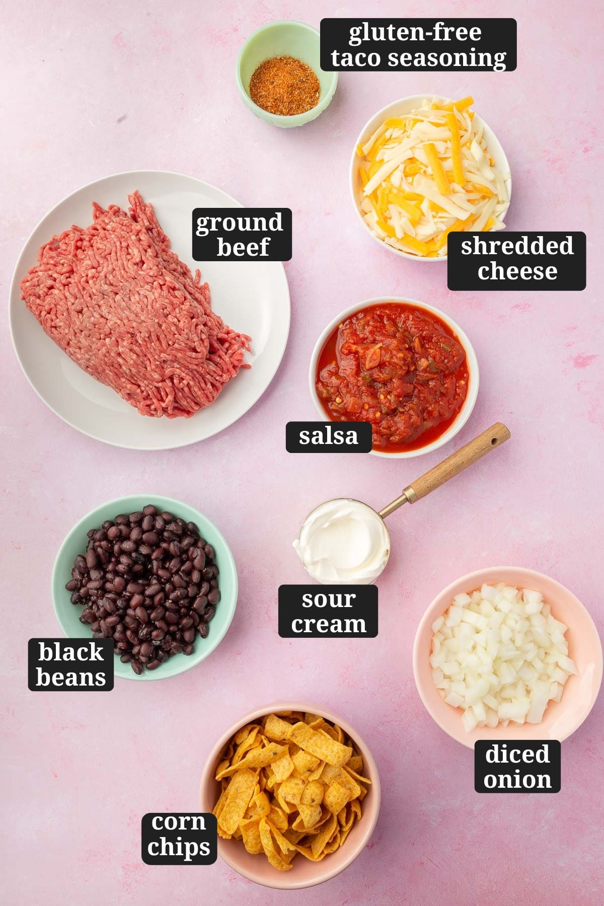 Ingredients in bowls to make walking taco casserole, including ground beef, gluten-free taco seasoning, shredded cheese, salsa, sour cream, black beans, Frito corn chips, and diced yellow onion with text overlays over each ingredient.