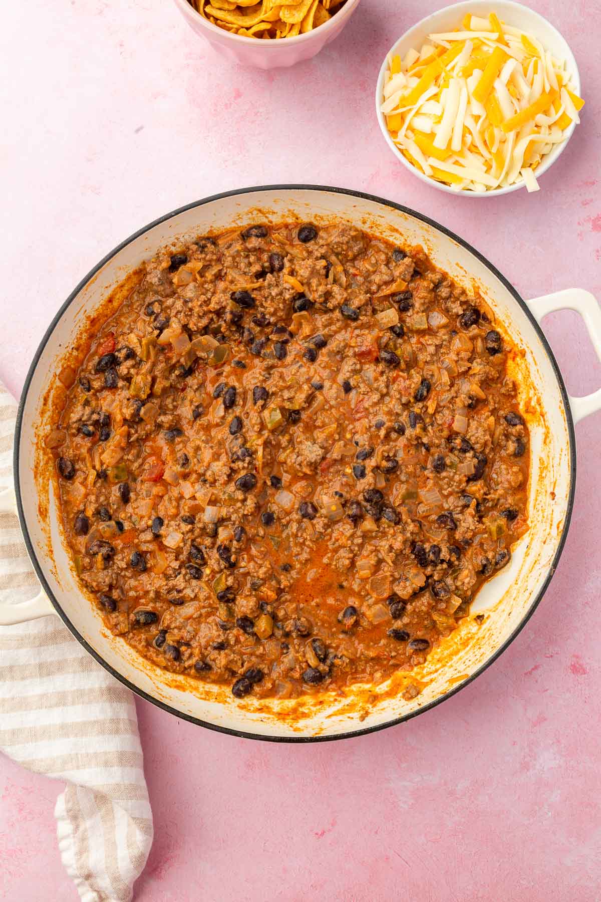 A white skillet with cooked ground beef, salsa, and black beans that have been simmered together with bowls of shredded cheese and corn chips to the side.