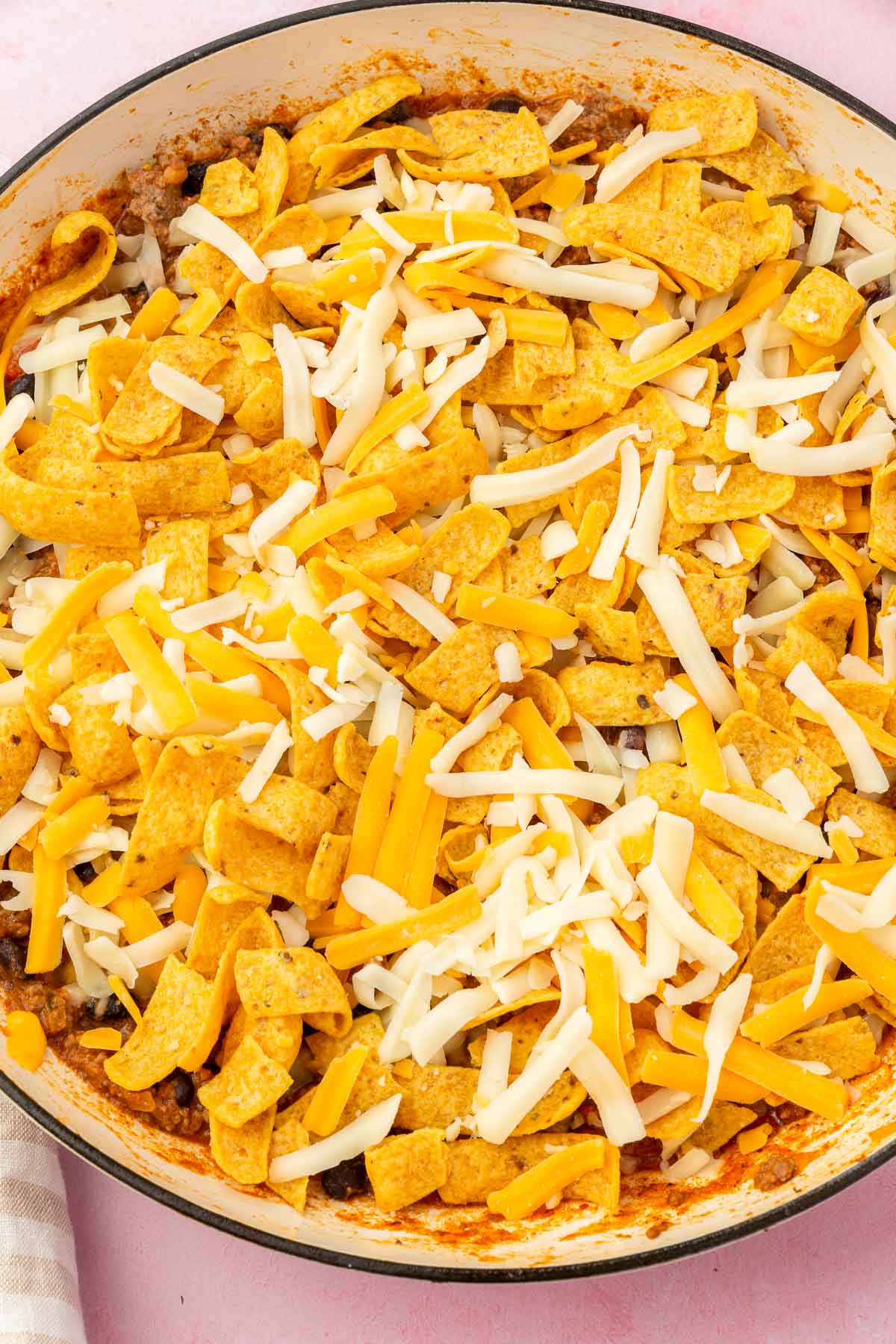 A skillet with ground beef topped with shredded cheese and Frito corn chips before baking in the oven.