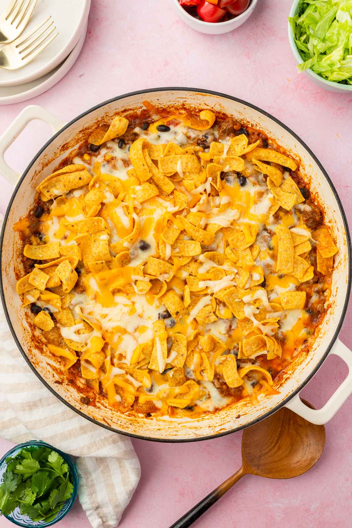 An overhead view of a skillet of walking taco casserole that has been baked until the cheese is melted with bowls of lettuce, tomatoes and cilantro to the side.