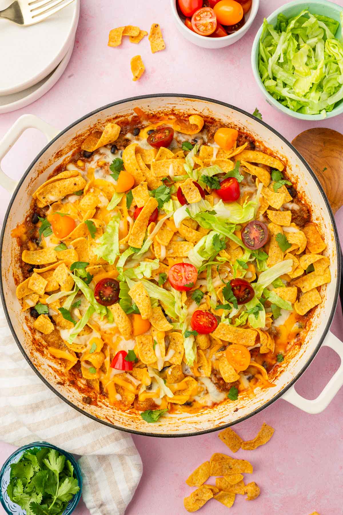 A braising pan of walking taco casserole topped with cherry tomatoes, lettuce and cilantro with bowls of toppings.