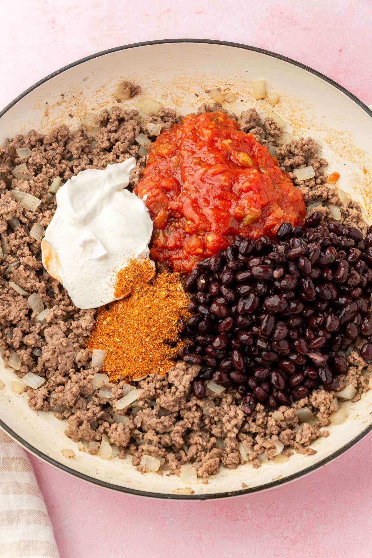 A skillet filled with cooked ground beef and topped with black beans, sour cream, salsa, and gluten-free taco seasoning before mixing together.