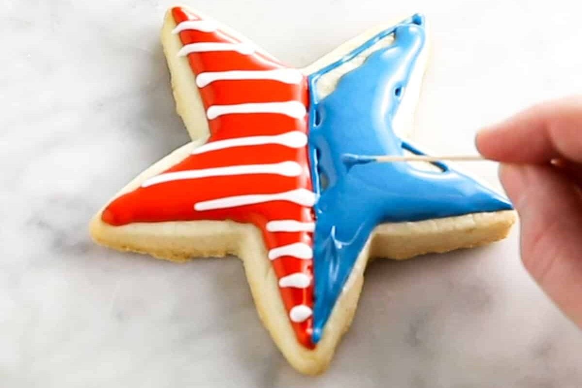 A star shaped sugar cookie that is being decorated with blue royal icing with a toothpick for 4th of July.