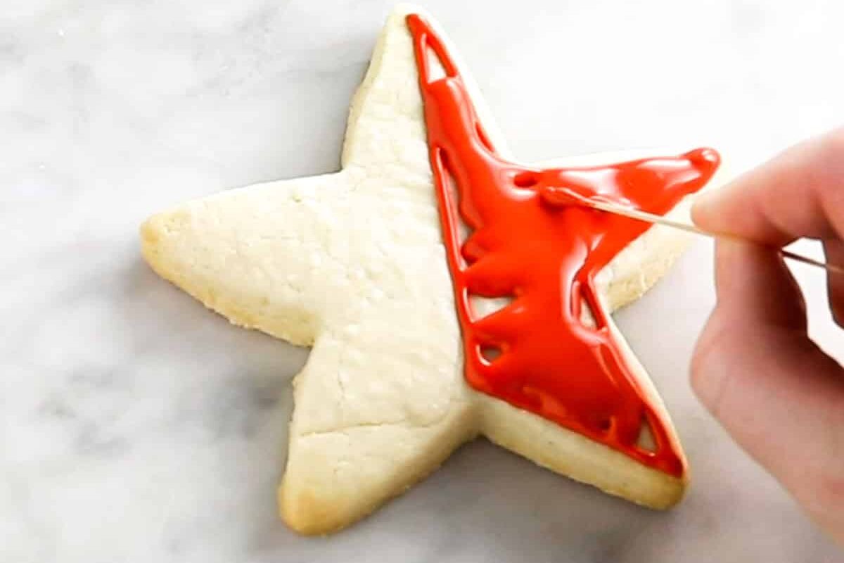 A star shaped sugar cookie that is being decorated with red royal icing and being spread with a toothpick.