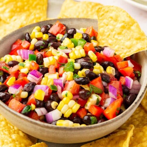A bowl of black bean corn salsa on top of a plate of tortilla chips with 3 appetizer plates stacked in the background.