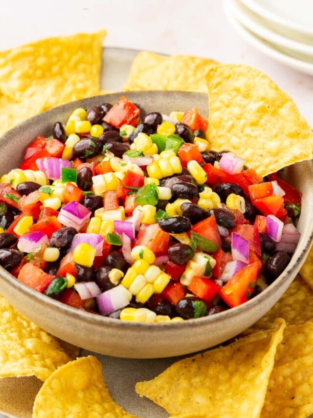 A bowl of black bean corn salsa on top of a plate of tortilla chips with 3 appetizer plates stacked in the background.