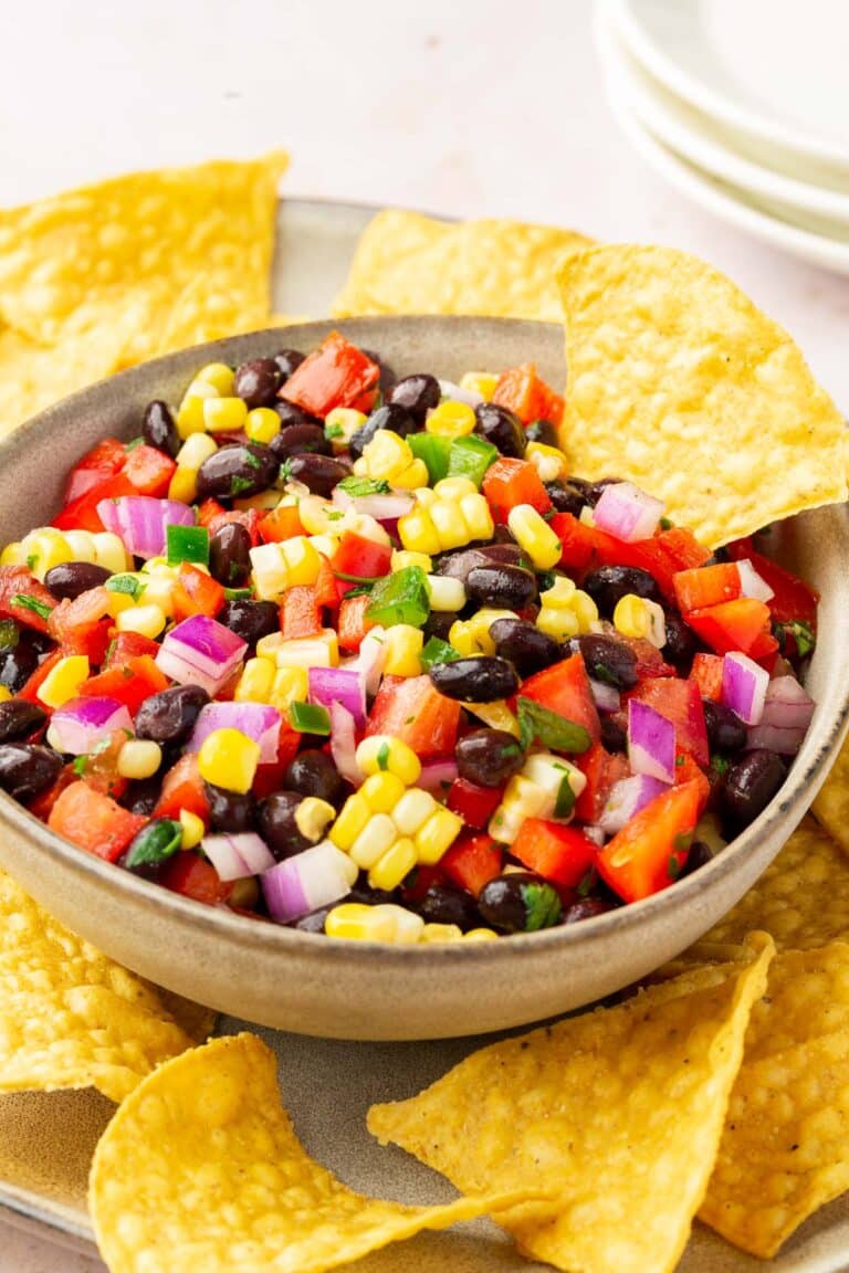 A bowl of black bean corn salsa on top of a plate of tortilla chips with 3 appetizer plates stacked in the background.