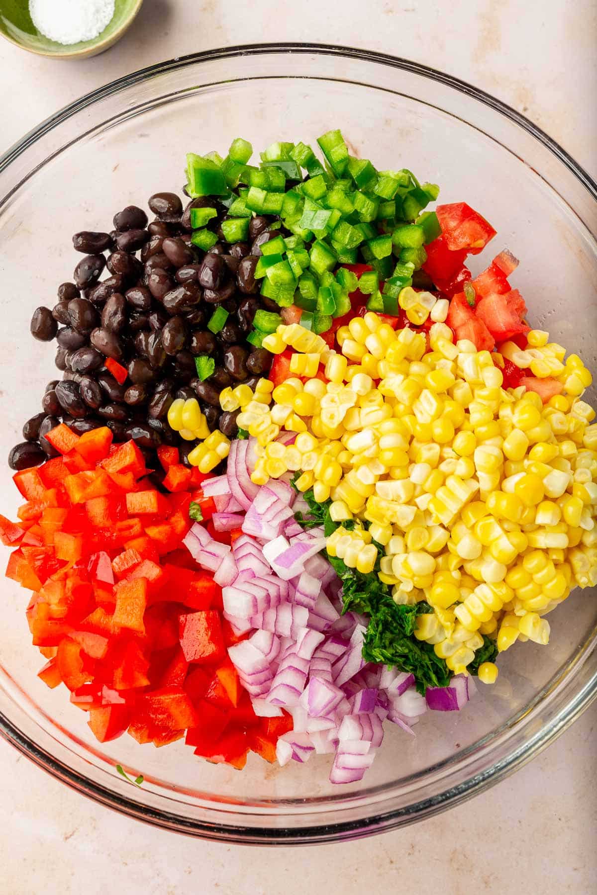 A glass mixing bowl with black beans, diced jalapeño, tomato, corn, cilantro, red onion and red bell pepper before mixing together.